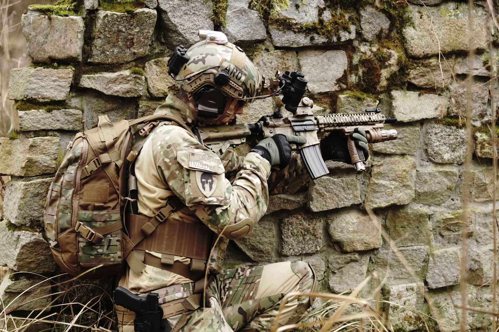 Soldier in camouflage gear aims a rifle while hiding along a stone wall outdoors.